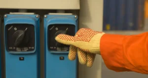 Mano de un trabajador con guante de seguridad y overol naranja manipulando uno de dos interruptores eléctricos azules en un tablero industrial